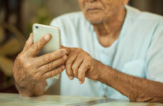 A senior man holding a smartphone in his right hand while tapping the screen using his left hand.