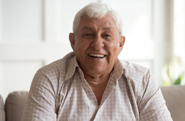 A senior man sitting on a couch smiling, looking directly at the camera