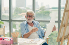 An image of a senior man holding a paintbrush in his right hand and a large piece of paper in his left hand as he smiles and looks at the camera.