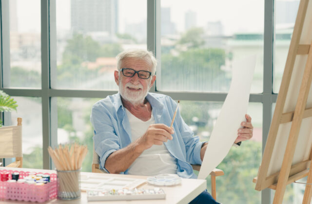 An image of a senior man holding a paintbrush in his right hand and a large piece of paper in his left hand as he smiles and looks at the camera.