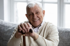 A senior man sitting on the couch smiling with both hands placed on his walking stick and looking directly at the camera
