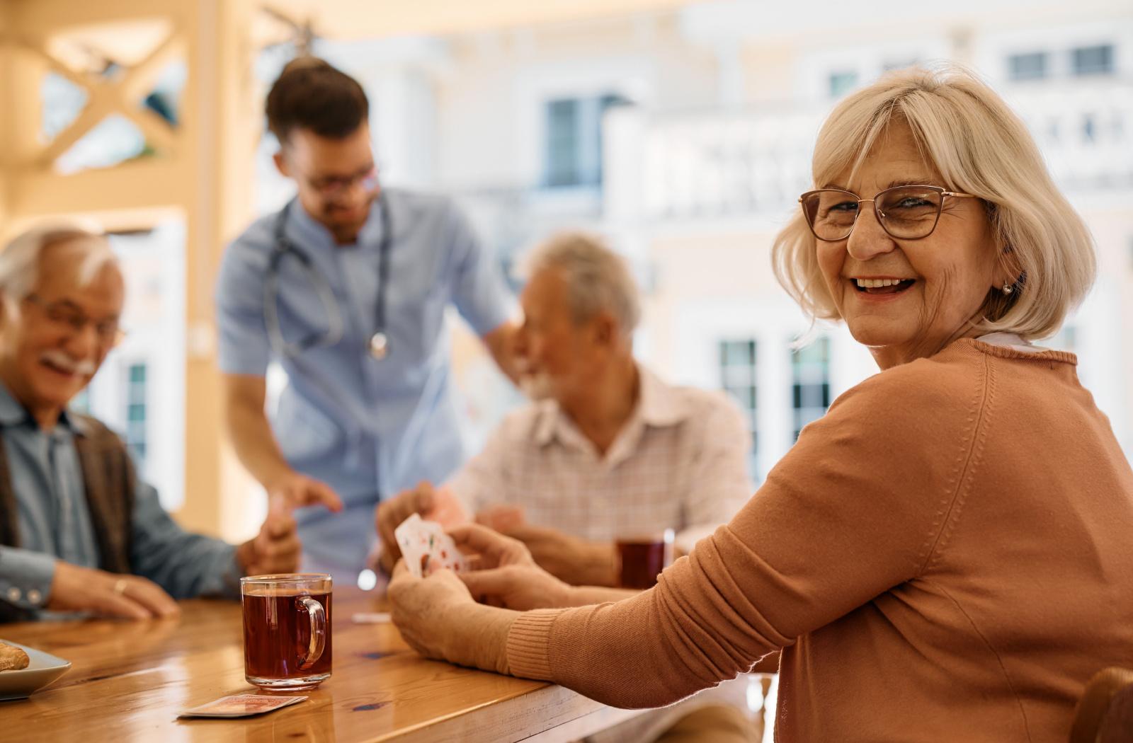 A group of older adults at a senior living community sit around a table laughing and playing cards together