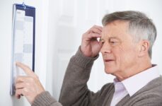 An older adult man with a forgetful look trying to remember something while pointing at a calendar.