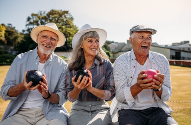 Three older adults smiling while holding lawn bowling balls outside.